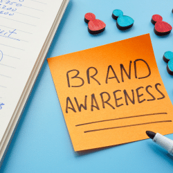 A post it note saying "Brand Awareness" on a blue desk next to a pad.