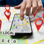 A bobile phone lays flat on a table with a person pressing a pin of a location on the screen. The phone is on a table made up of a digital map with 3d pins all around. There is a "Local SEO" title on white wooden blocks in the bottom left hand corner.