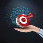 A hand holding a target with a dart in the bullseye with a cloud of holographic email, money, and charts surrenounding the target.