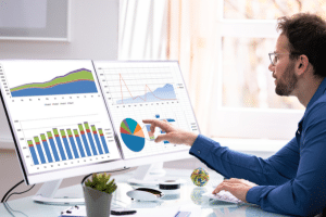 Businessman sitting at a modern desk with 2 screens comparing analytics data.
