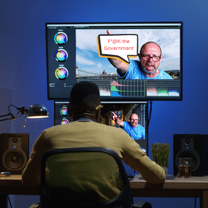 A young man sitting at his dual screen editing setup adding expletives to a video he's changed the background on.