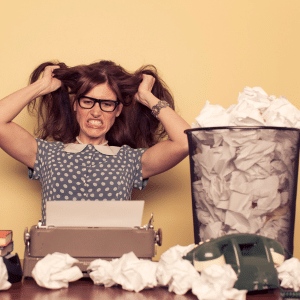 Lady sat at a typewriter pulling her hair out next to a bin full of screwed up paper.
