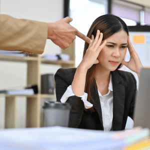 Arm of a man pointing a finger at a young professional lady sat at her desk looking very stressed.
